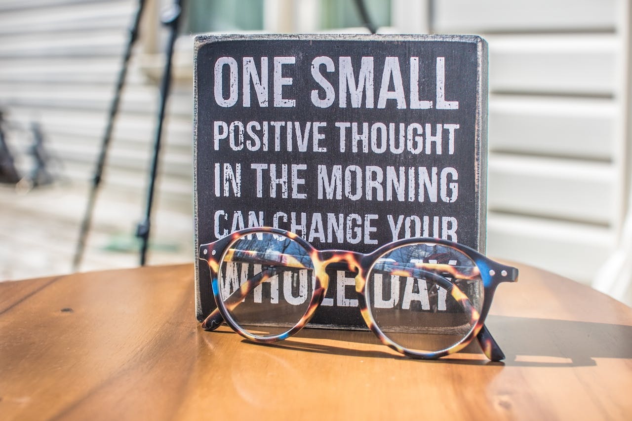 gallery-3 Eyeglasses resting on a table in front of a motivational quote sign, emphasizing positivity.